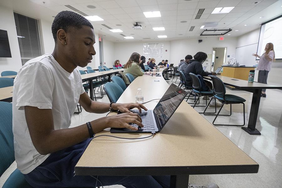 A student works on their laptop as a lecturer stands in the front of class.